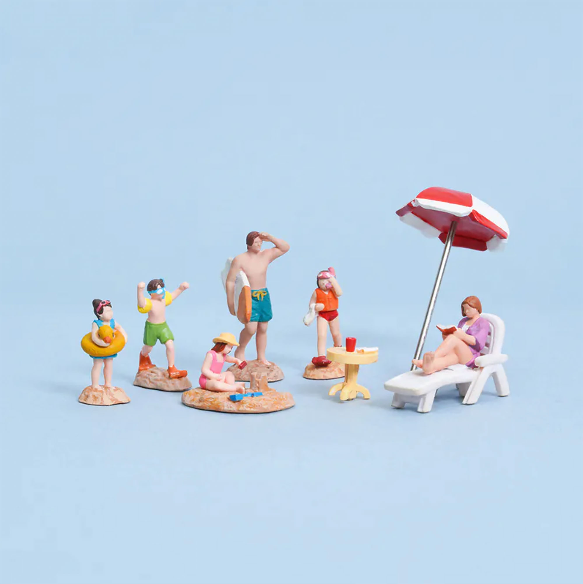 Miniature Beach People and Decor Figurines: A collection of miniature resin figurines and accessories depicting a beach scene (scène de plage miniature, Miniatur-Strandszene, escena de playa en miniatura). There are figures of people in swimwear (maillots de bain, Badeanzüge, trajes de baño): one holding a surfboard (planche de surf, Surfbrett, tabla de surf), one with snorkeling gear (équipement de plongée, Schnorchelausrüstung, equipo de snorkel), one walking, and one holding a swim ring (bouée de natation, Schwimmring, flotador). Below, there’s a figure lounging on a beach chair (chaise longue, Liegestuhl, tumbona) reading a book, another building a sandcastle (château de sable, Sandburg, castillo de arena), and a small table with a drink under a red and white beach umbrella (parasol, Sonnenschirm, sombrilla).